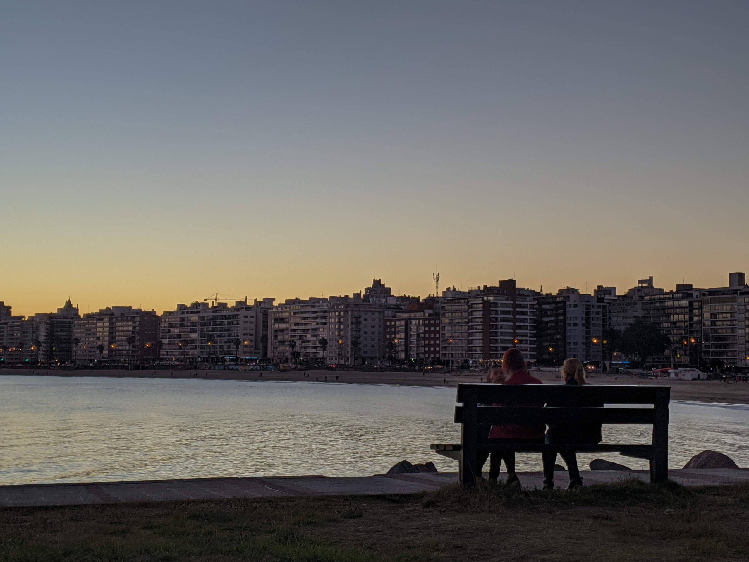 Pessoas sentadas em um banco de frente para o mar em Montevideo, aos fundos é possível ver alguns prédios e o dia está se pondo.