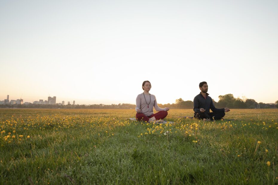 Casal sentado em posição de meditação em um bosque, ao fundo é possível ver uma cidade.