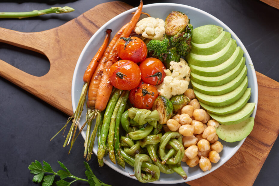 Foto de um prato de comida sobre uma tabua de madeira, nele estão diversos vegetais, frutas e grãos: cenoura, abacate, tomate, grão de bico, vagem e quiabo.