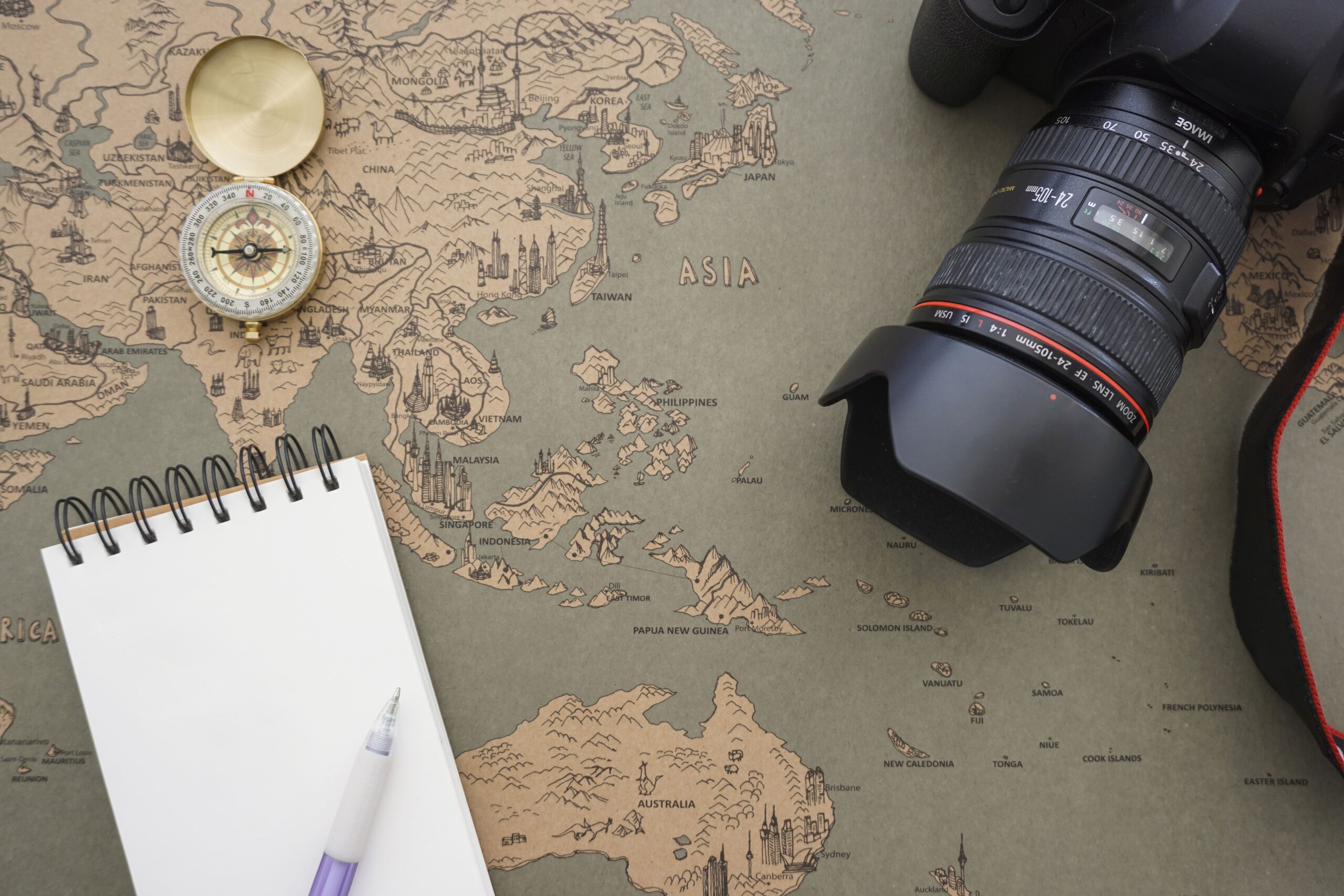 Foto de uma mapa visto de cima, em cima dele está um bloco de notas, uma caneta, uma bússola e uma câmera, representando os documentários de viagem.