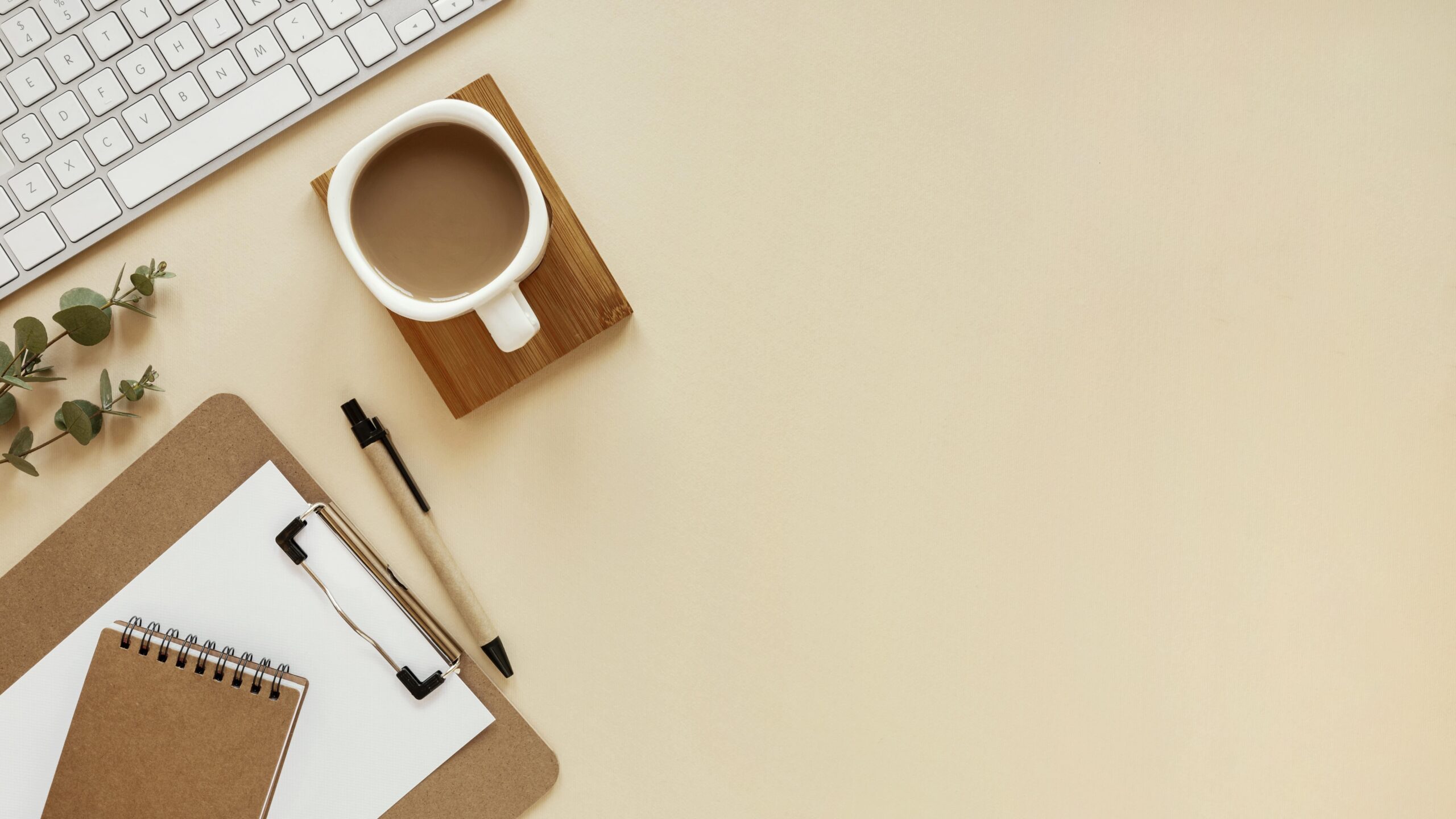 Foto de uma mesa de trabalho vista de cima, como uma xícara de café, caneta, prancheta com pepeis e um bloco de notas em cima, o teclado de um notebook e algumas folhas, representando um ambiente agradável e a saúde mental do trabalhador que é impactada pela jornada de trabalho de 4 dias.