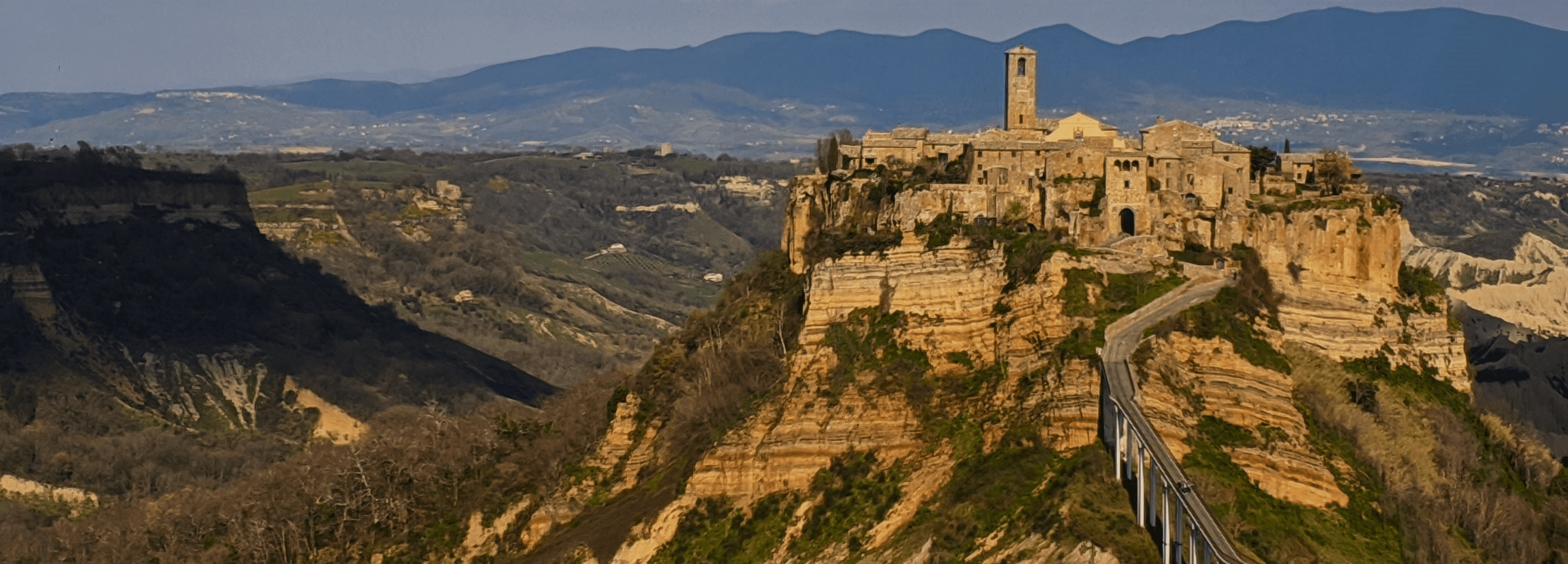 Foto de Civita di bagnoregio vista de longe, é possível ver suas casas no topo da montanha no estilo medieval, com uma ponte que leva até ela.