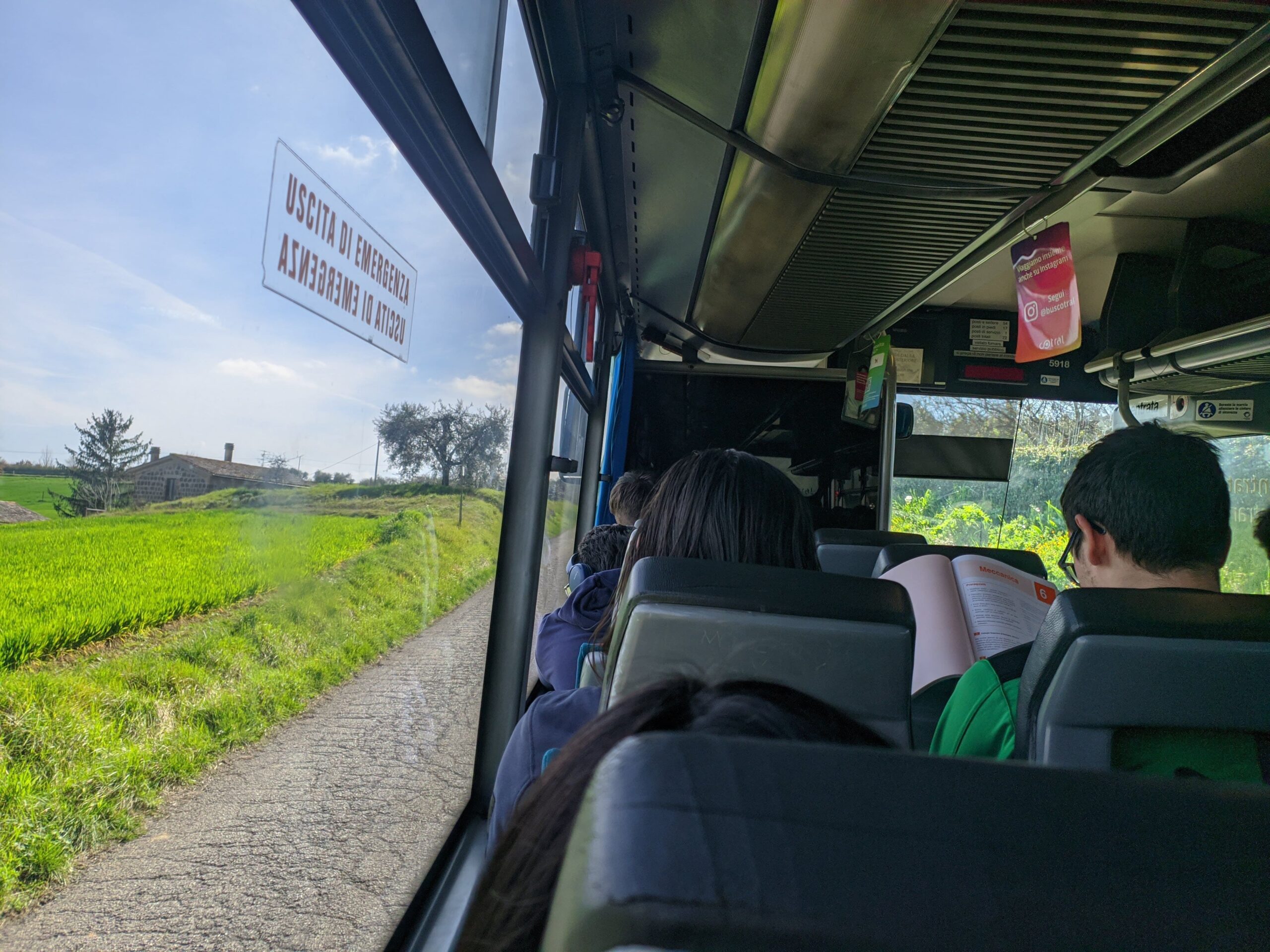 Foto tirada dentro de um ônibus, é possível ver algumas pessoas sentadas, uma lendo, e a paisagem verde para fora.