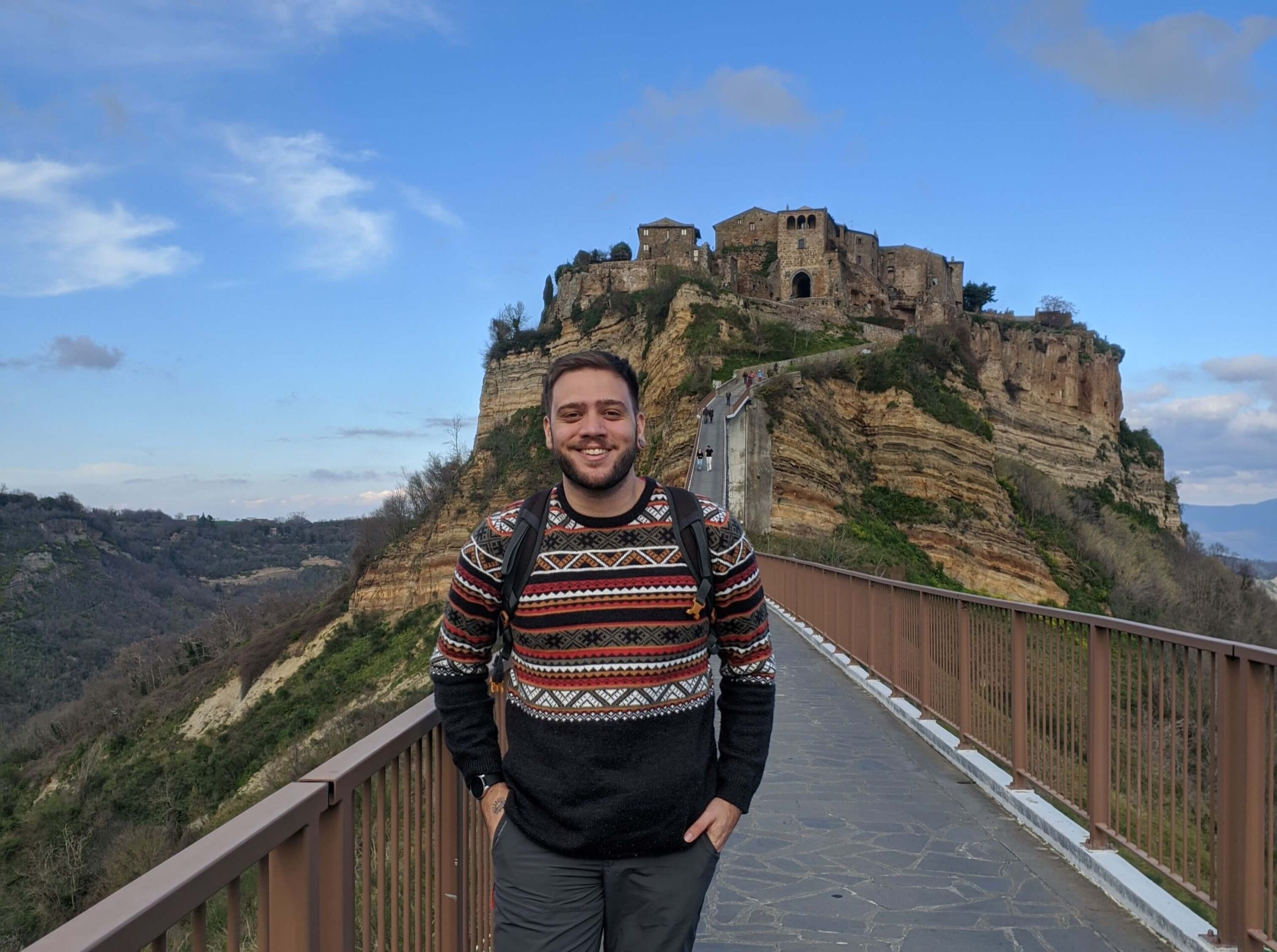 homem vestido com casaco com padrões étnicos e sorrindo na ponte de civita di bagnoregio, com a cidade ao fundo