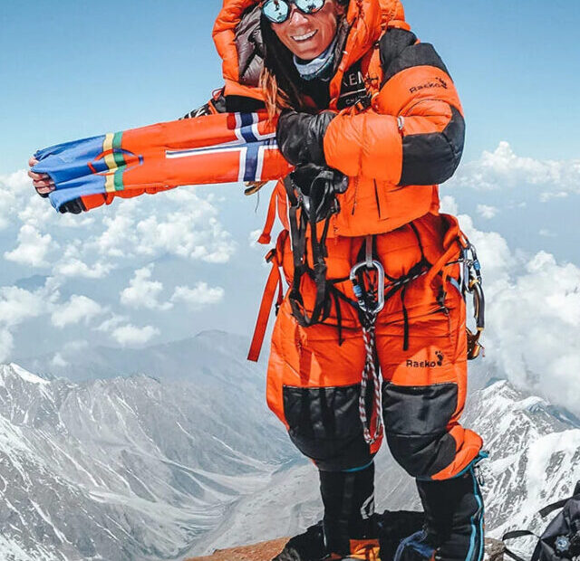 Foto de Kristin Harila no cume de uma montanha, é possível ver nuvens e outros picos atrás dela, a montanhista está de pé com uma roupa laranja grossa que cobre o seu corpo enquanto sorri.