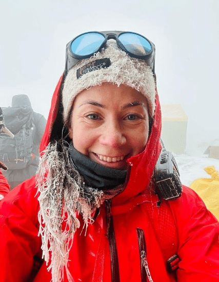 Selfie de Kristin Harila, ela está sorrindo para a foto, usando um gorro branco, óculos escuros pendurados na cabeça e um casaco laranja.