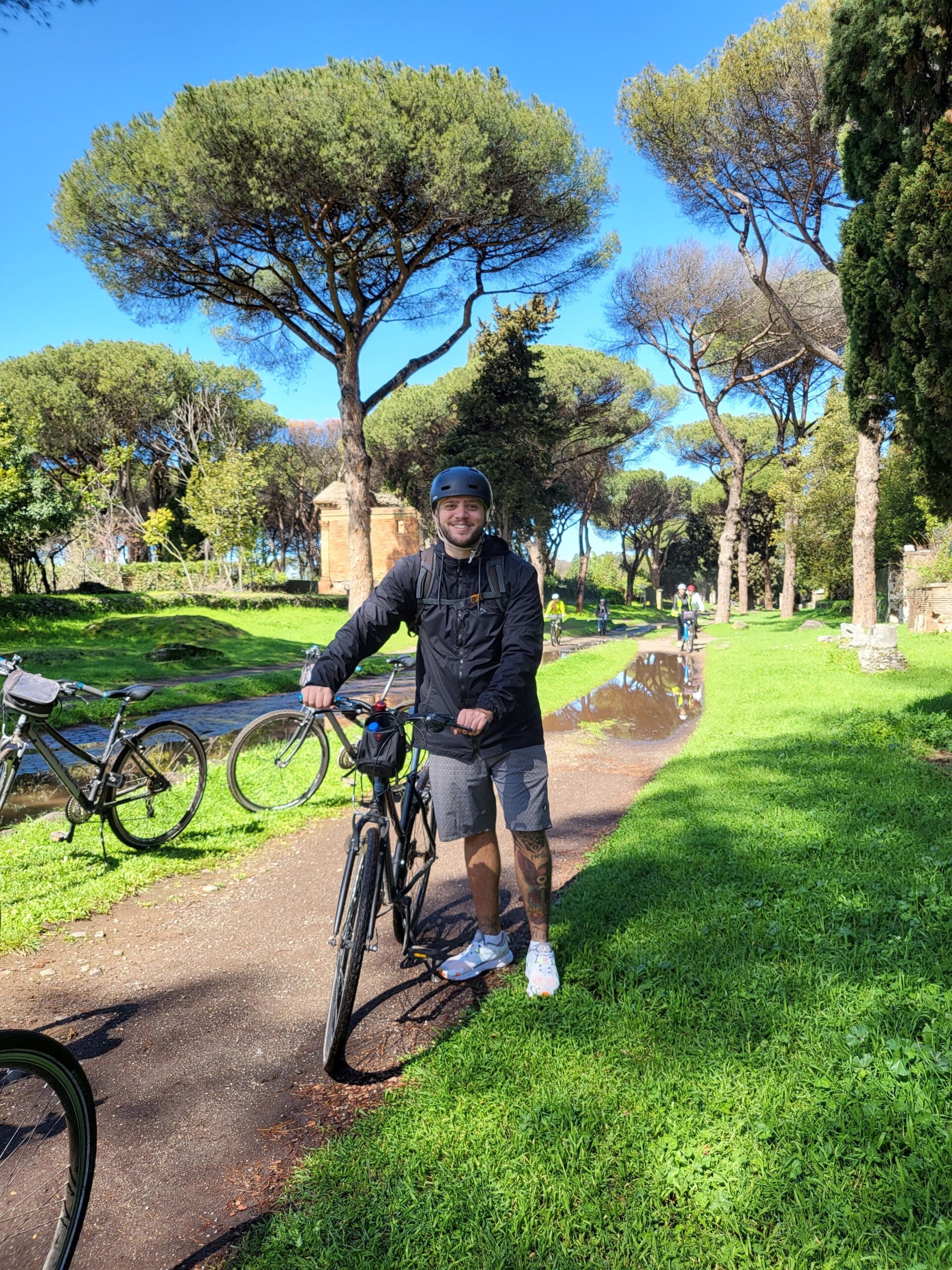 Foto minha na Via Ápia, segurando uma bicicleta com a estrada de fundo e sorrindo para a foto, de capacete.