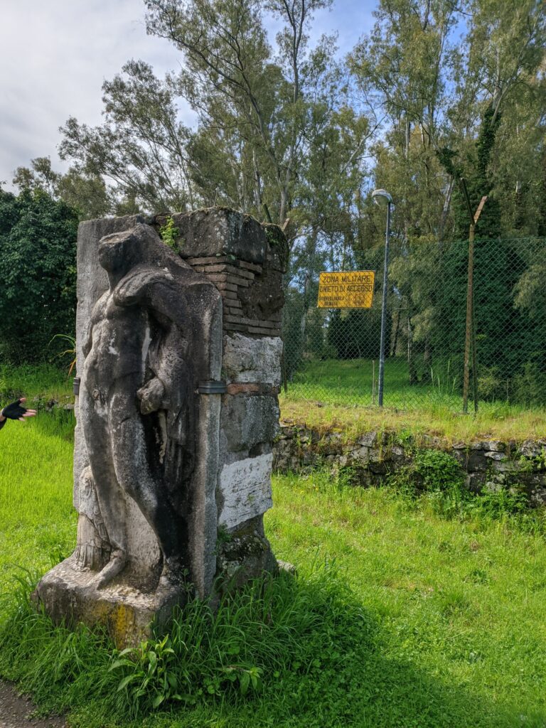 Foto de uma estátua sem a cabela e partes do seu corpo, exposta em um gramado e sustentada por uma estrutura de pedras e tijolos.