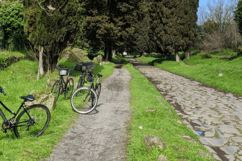 Foto de bicicletas estacionadas ao lado via ápia, que é feita de pedras e com gramas e árvores ao redor.