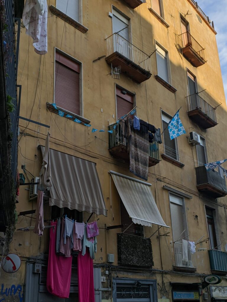 Foto da fachada de um prédio em Nápoles, sua cor é amarelo-escuro, é possível ver toldos antigos em cima das janelas, algumas roupas penduradas logo em baixo deles e em algumas janela há bandeiras azuis.