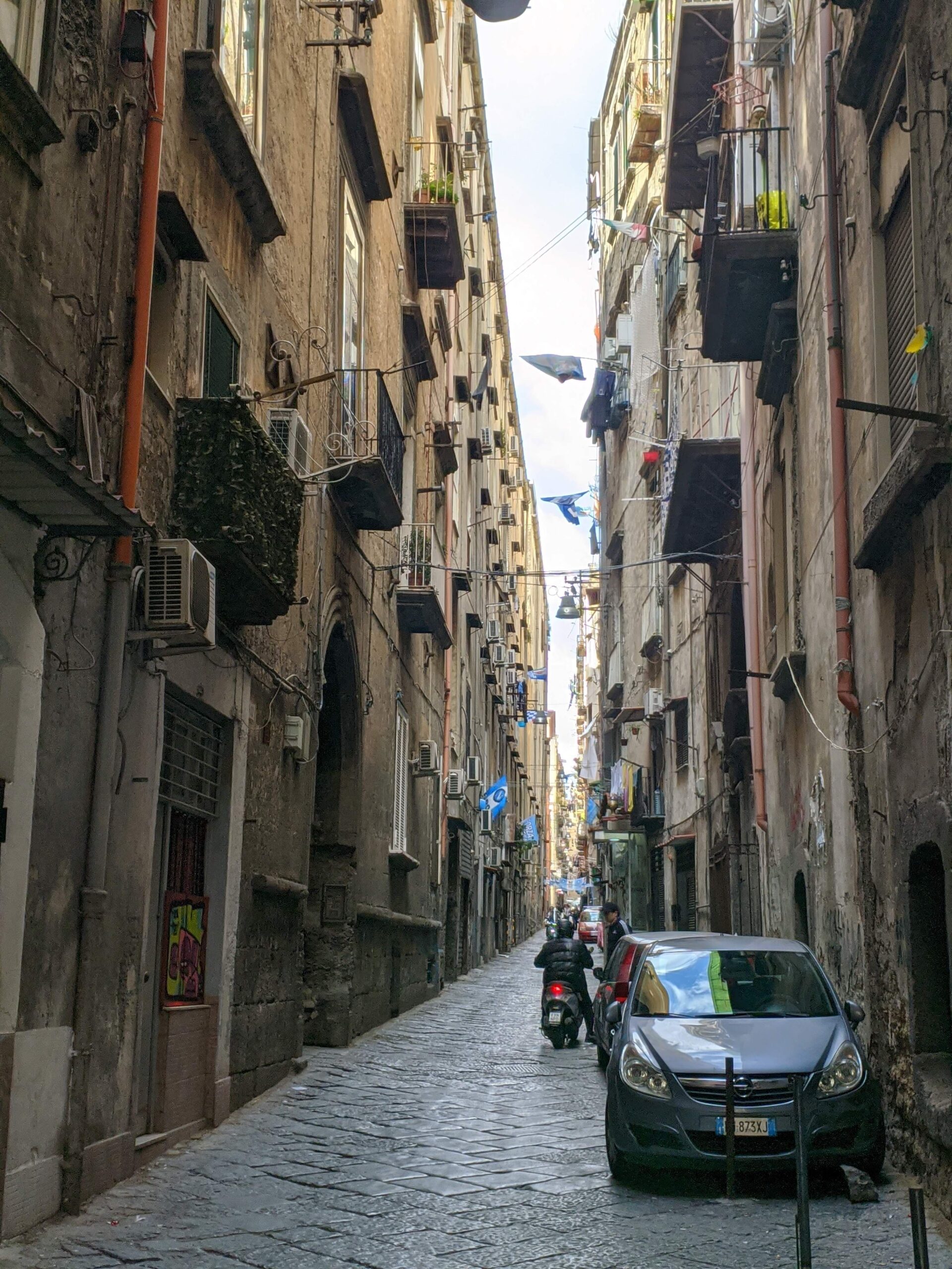 Foto de uma rua de Nápoles, ela é estreita, com prédios de altura mediana e cor escura dos dois lados, que também acabam escurecendo as rua.