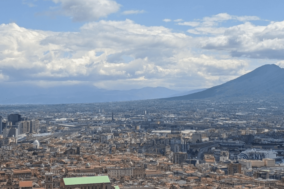 Foto de Nápoles vista de cima, é possível ver diversos prédios e ao fundo o Vesúvio, em formato de "corcova de camelo"