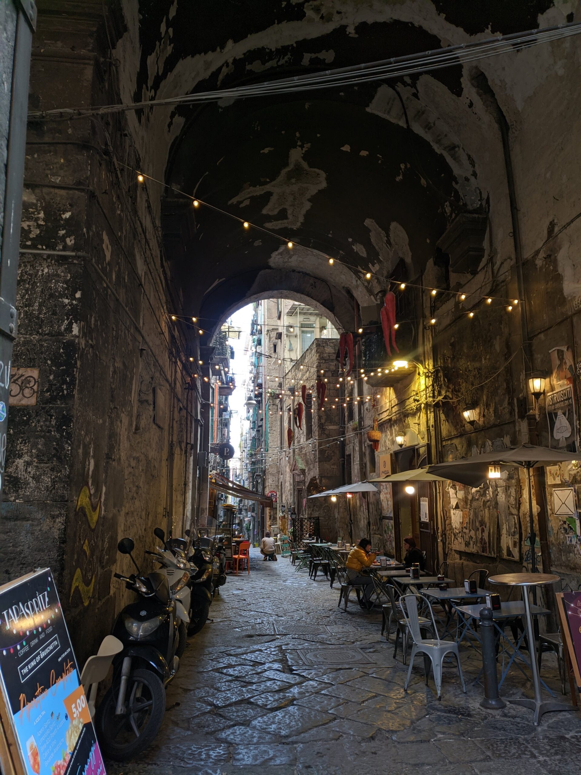 Foto de uma galeria formada por um arco em Nápoles, nela é possível ver um varal de luzes, mesas de um restaurante e várias motos encostadas em sua parede.