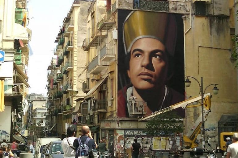 Foto de uma rua com prédios, em um dele está um mural que retrata uma pessoa olhando para cima com roupas eclesiásticas. 