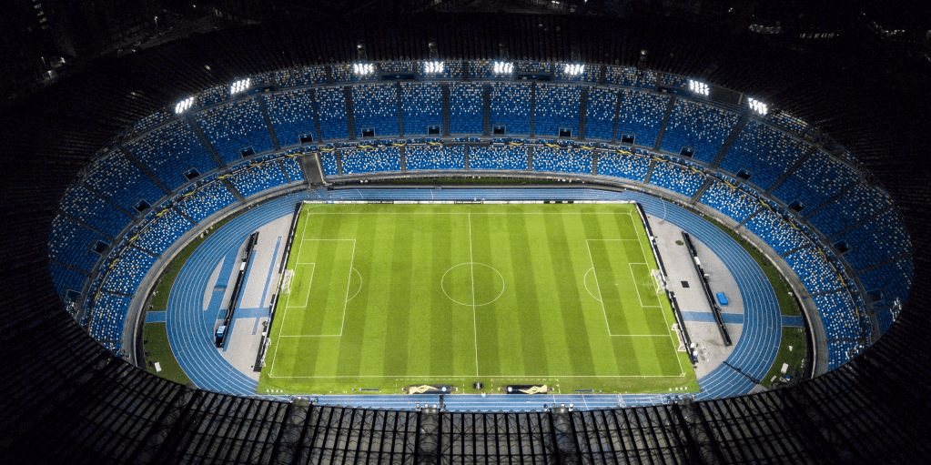 Foto de um estádio visto de cima, com o gramado verde e arquibancada de dois andares em diferente tons de azul.