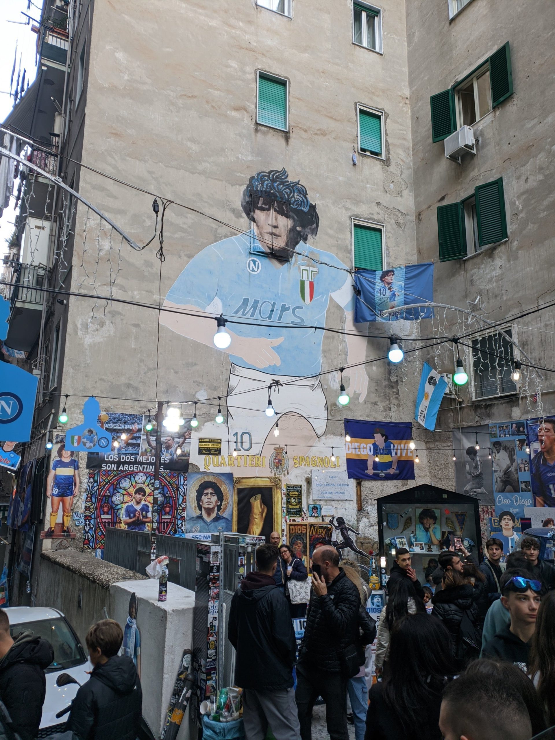 Foto da fachada de um prédio, nele está um mural que retrata Maradona, de uniforme do Napoli como se estivesse jogando futebol.