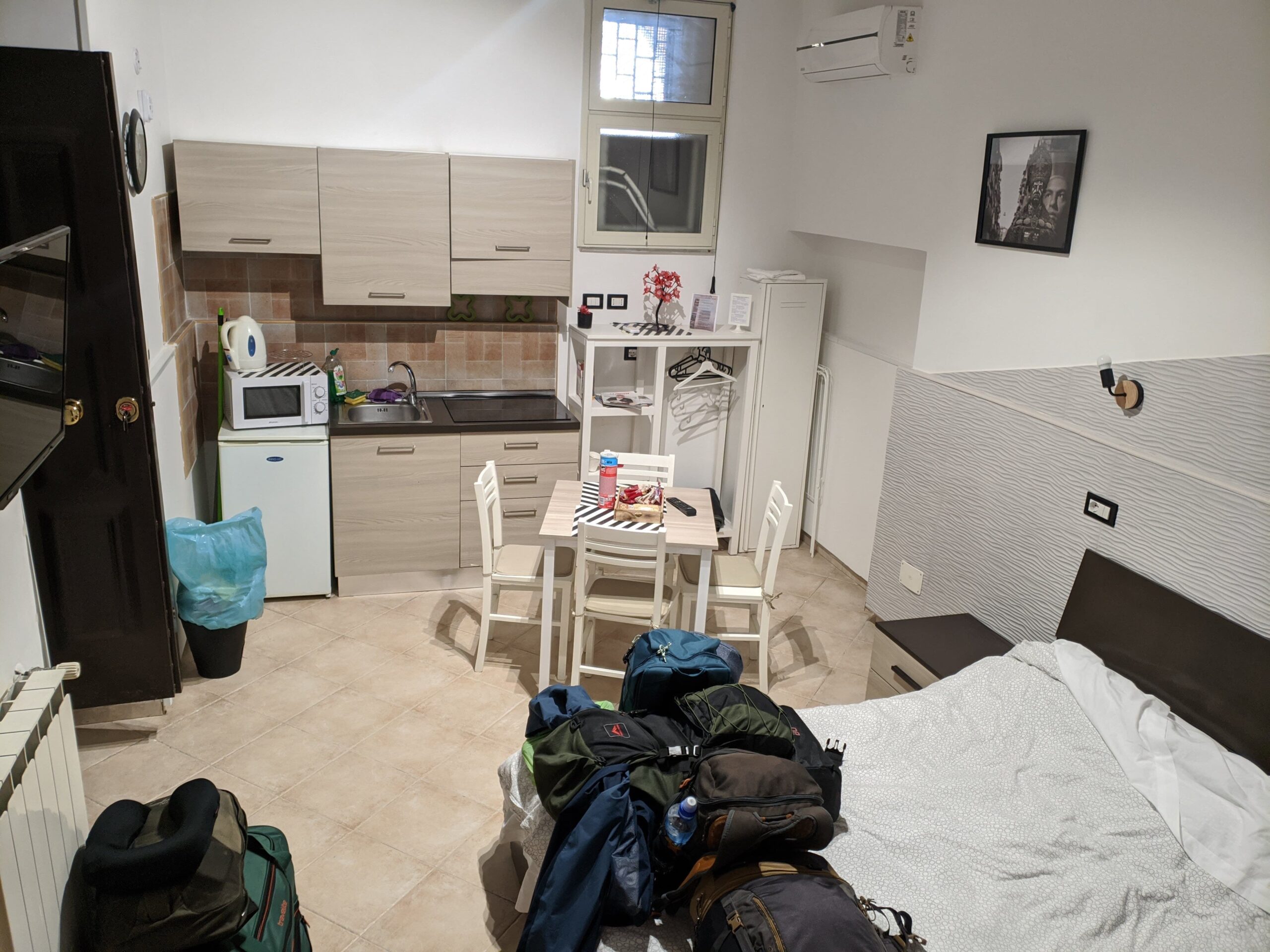 Foto de um quarto, com uma cama, uma pequena cozinha ao lado, uma mesa ao lado da cama e em frente a cozinha e mochilhas em cima da cama.