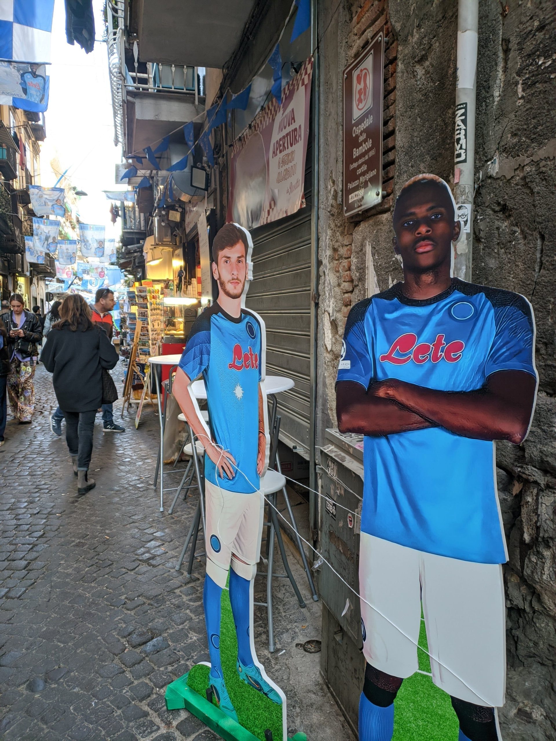 Foto de duas placas que representam um homem cada em meio a uma rua de Nápoles, ambos estão com o uniforme de Napoli e são jogadores do time.
