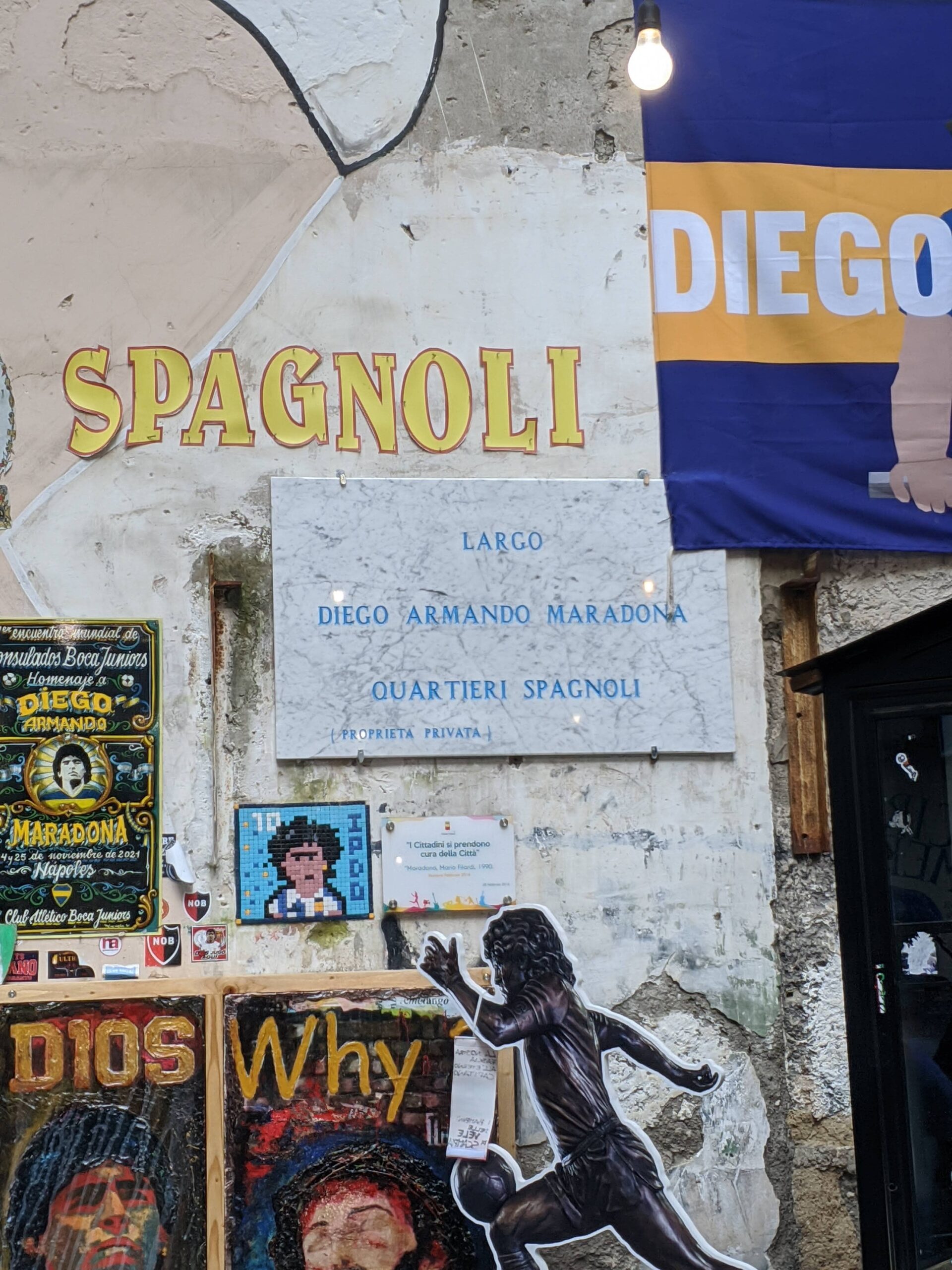 Foto de uma placa de mármore colada em um parede que diz: Largo Diego Maradona, Quartieri Spagnoli, local onde fica o "santuário" de Maradona em Nápoles.
