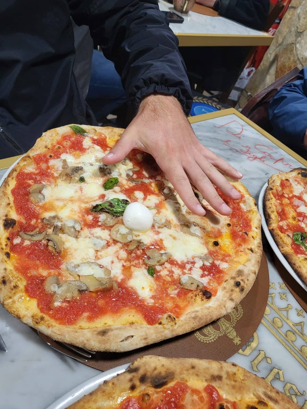 Foto de uma pizza com uma mão em cima, para mostrar o seu tamanho, a pizza da cerca de 3 vezes o tamanho da mão.