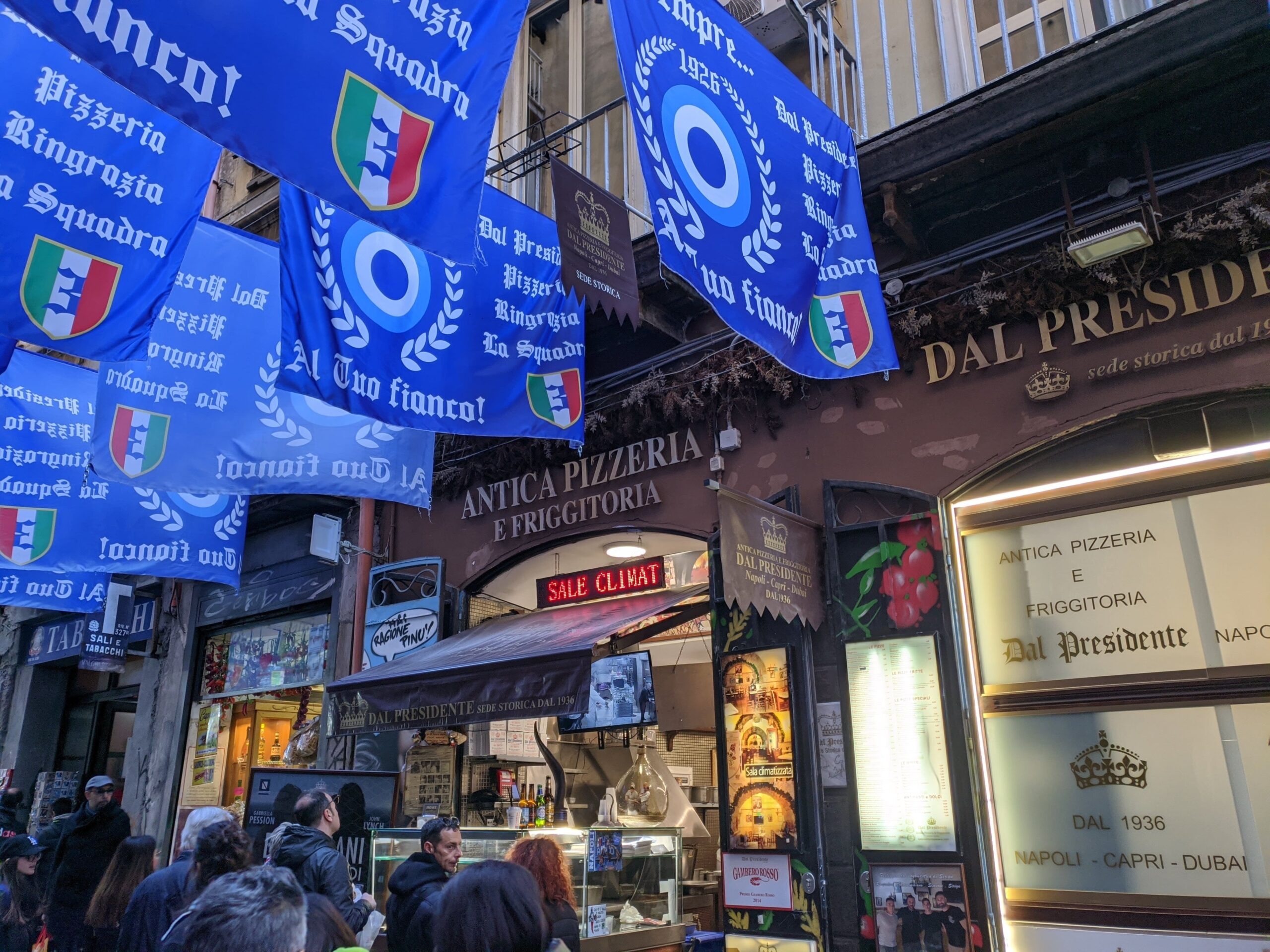 Fachada de uma pizzaria, com bandeiras azuis remetendo ao Napoli em cima dela, pessoas a sua frente e o letreiro com o seu nome em cima da porta "Pizzeria dal presidente"