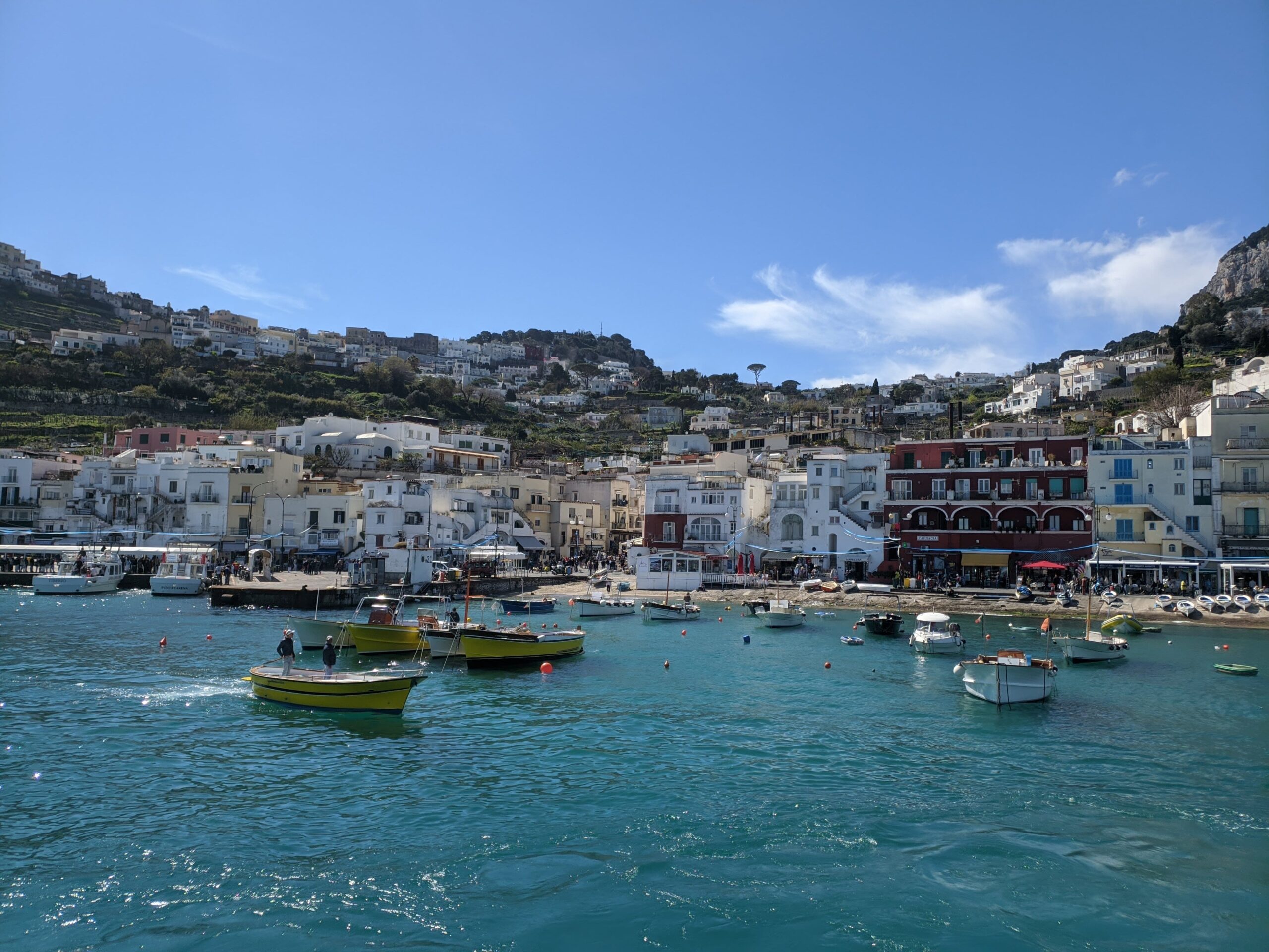 Foto de Capri vista a partir do mar, é possível ver a cidade com casas predominantes brancas atrás da praia e que sobem a encosta no entorno. O mar está azul esmeralda e com alguns barcos navegando.