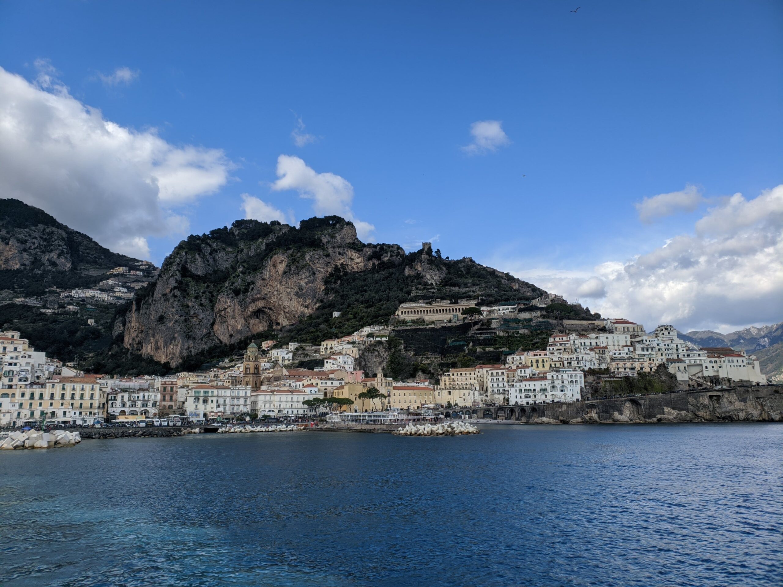 Foto de Amalfi vista do mar. É possível ver uma grande montanha rochosa com casas de tons branco amarelado que sobem até a sua metade. O mar está azul.