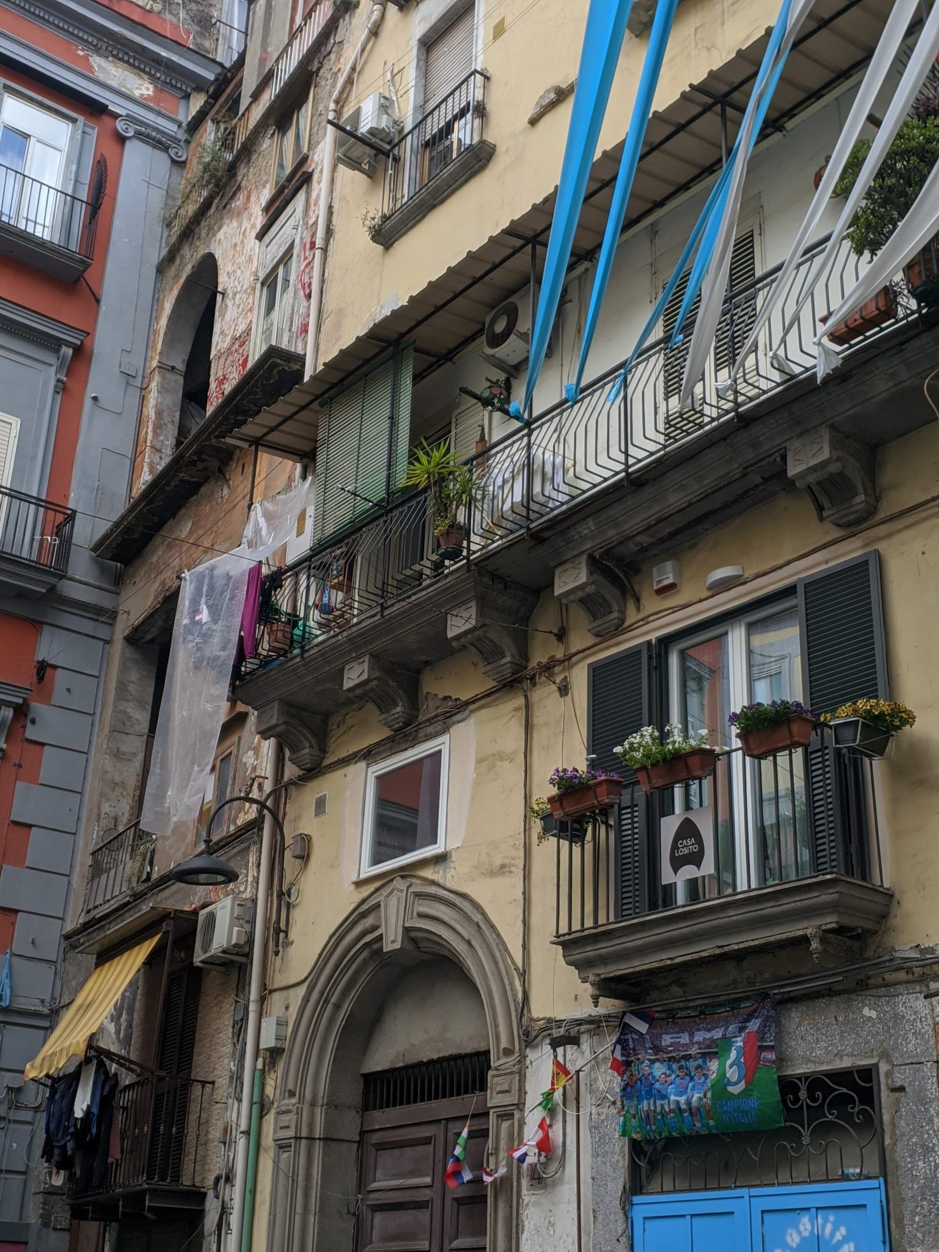 Foto da fachada de um prédio em Nápoles, o edifício é no tom amarelado e possui vários elementos, como janelas adornadas com plantas , uma porta contornada por um pórtico de pedra, faixas azuis e brancas que remetem ao time da cidade e roupas penduradas nas janelas.