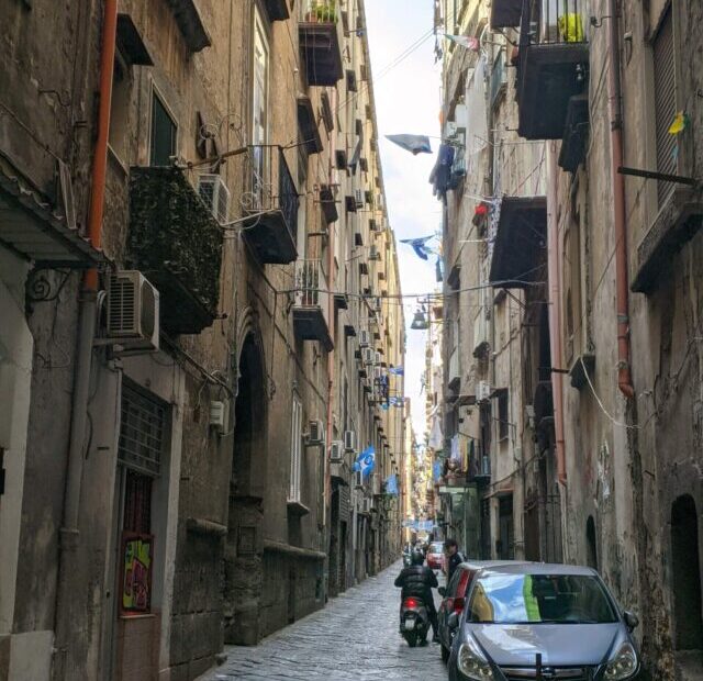 Foto de uma rua de Nápoles, ela é estreita, com prédios de altura mediana e cor escura dos dois lados, que também acabam escurecendo as rua.