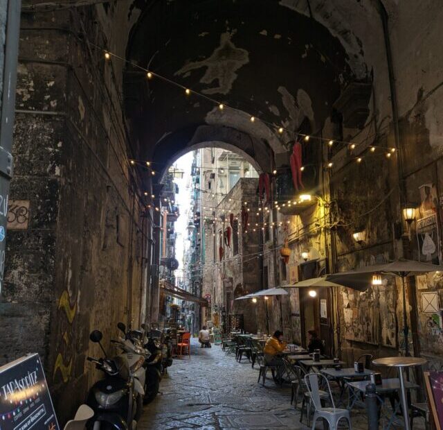 Foto de uma galeria formada por um arco em Nápoles, nela é possível ver um varal de luzes, mesas de um restaurante e várias motos encostadas em sua parede.