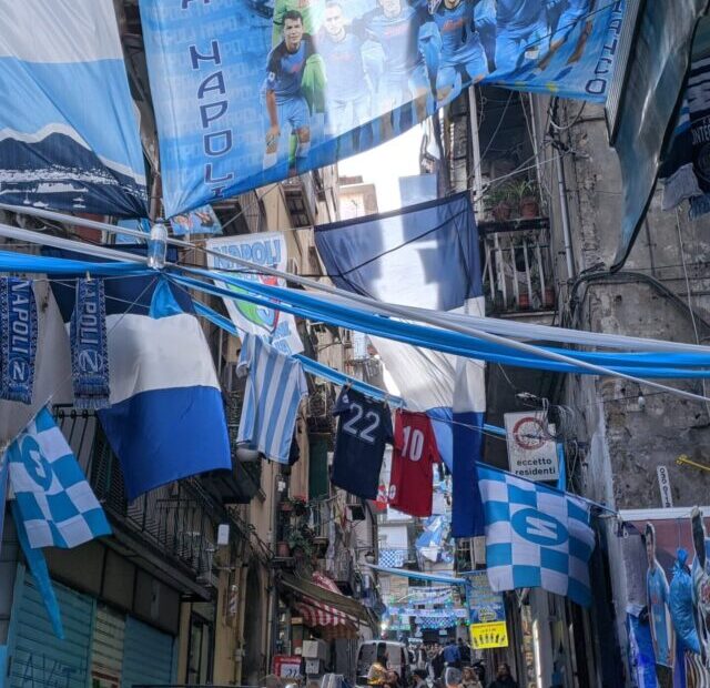 Uma rua com prédio antigos repleta de bandeiras e faixas nas cores azul e branca que cruzam de um prédio para outro.