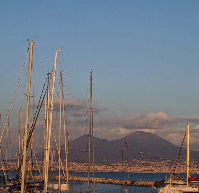 Foto do monte Vesúvio visto a partir da linha do mar. A montanha em formato de corcova de camelo está ao fundo com algumas nuvens sobre ela. A frente está o mar e vários valeiros parados.