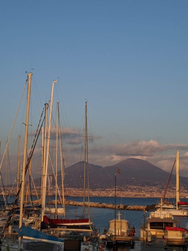 Foto do monte Vesúvio visto a partir da linha do mar. A montanha em formato de corcova de camelo está ao fundo com algumas nuvens sobre ela. A frente está o mar e vários valeiros parados.