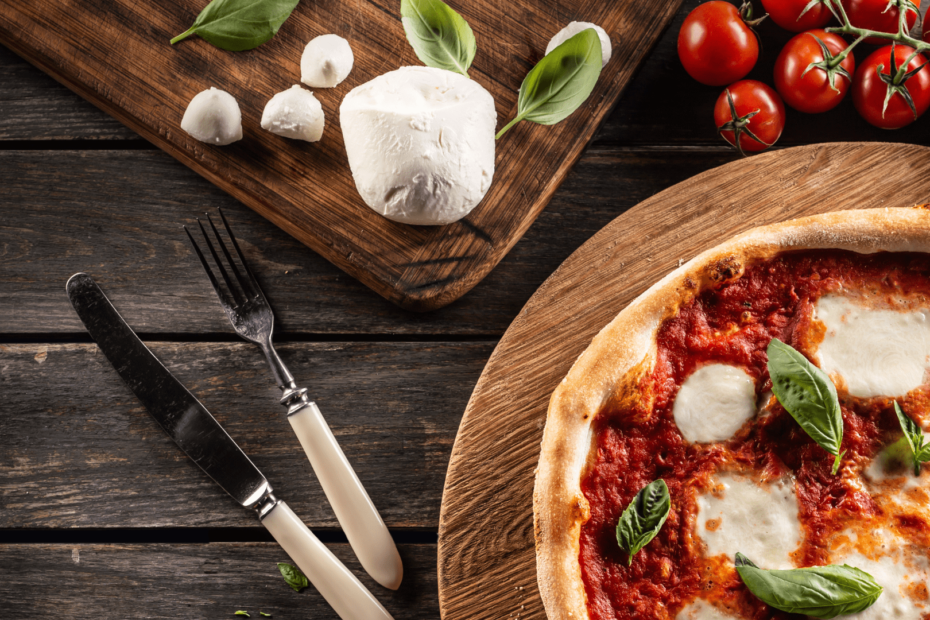 Foto de uma pizza vista de cima, é possível ver o seu molho vermelho, o queijo branco e algumas folhas de manjericão verde. Ela está sobre uma mesa de madeira marrom escura, ao seu lado está uma faca, uma tabua com queijo branco e alguns tomares-cereja vermelhos.