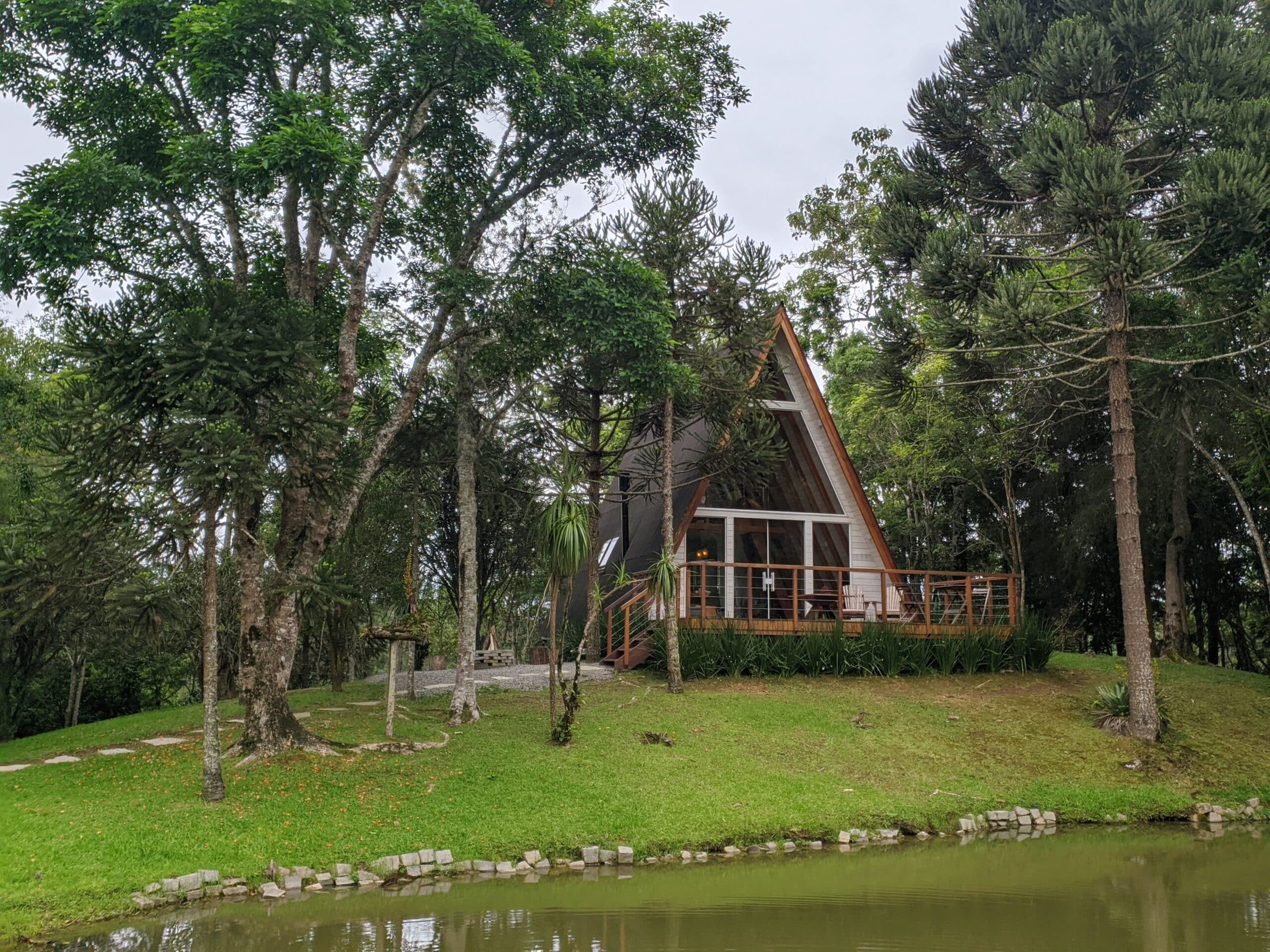 Imagem externa de uma cabana perto de Curitiba com teto em forma de A, situada em um gramado verdejante com árvores ao redor e à beira de um pequeno lago. O céu está nublado, e a cabana de madeira se integra à paisagem natural.