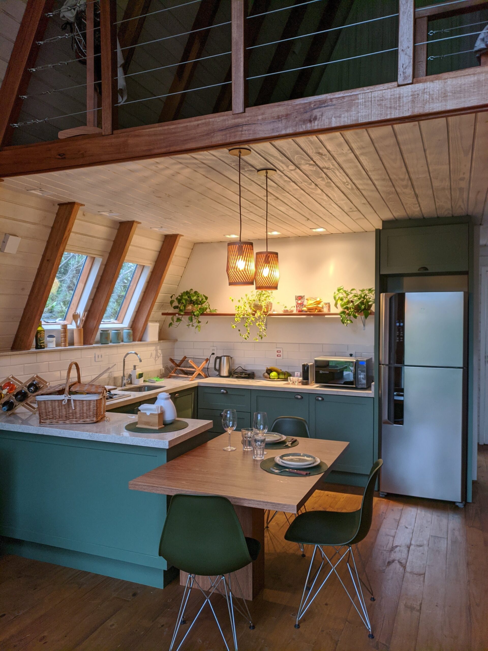 Uma cozinha de cabana com design interior moderno, caracterizada por armários em tom verde-escuro e balcão de pedra clara. Acima, pendem luminárias elegantes de madeira. O espaço é iluminado por luz natural proveniente das janelas triangulares no teto de madeira.