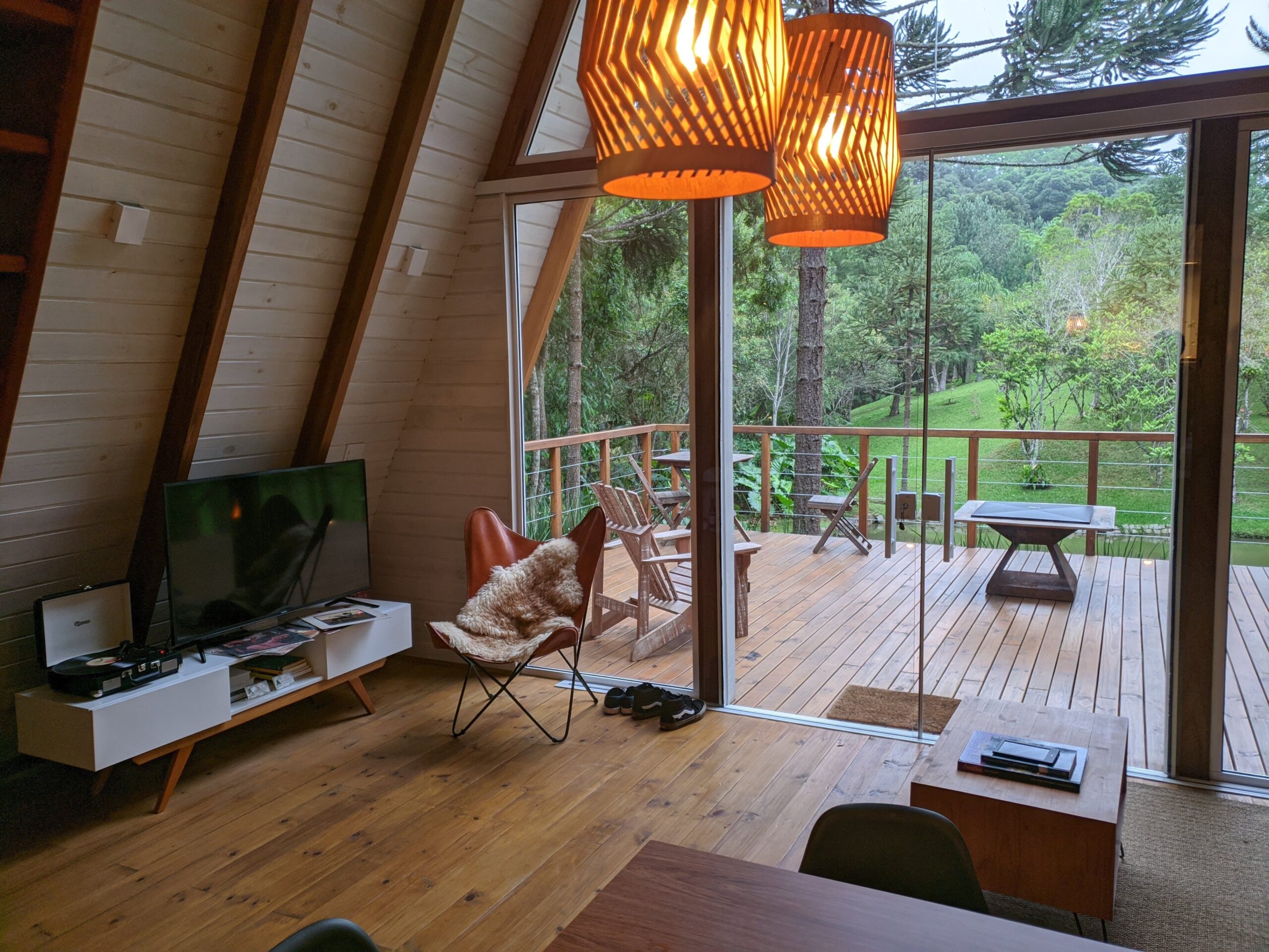 Interior de uma sala de estar em uma cabana, com teto de madeira em forma de A e grandes janelas de vidro que revelam uma varanda de madeira e uma vista verdejante para uma floresta. Há uma televisão, um sofá de couro marrom com uma pele fofa e uma mesinha de centro com livros.