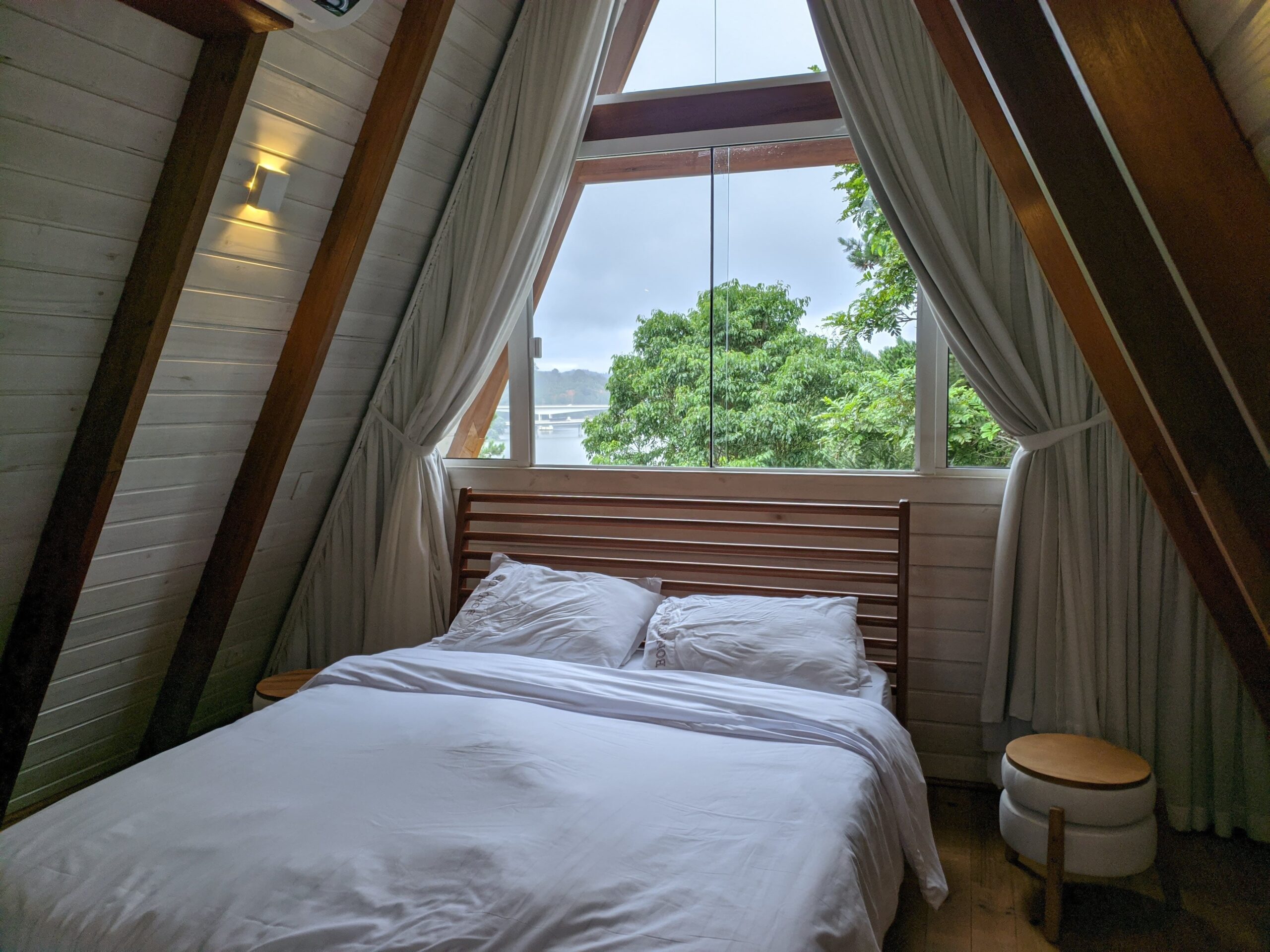 Quarto de uma cabana com grande janela triangular e uma cama de casal com lençóis brancos. O quarto é iluminado por uma luz suave e rodeado por cortinas brancas, criando um ambiente aconchegante. O exterior visível através da janela mostra um dia nublado.