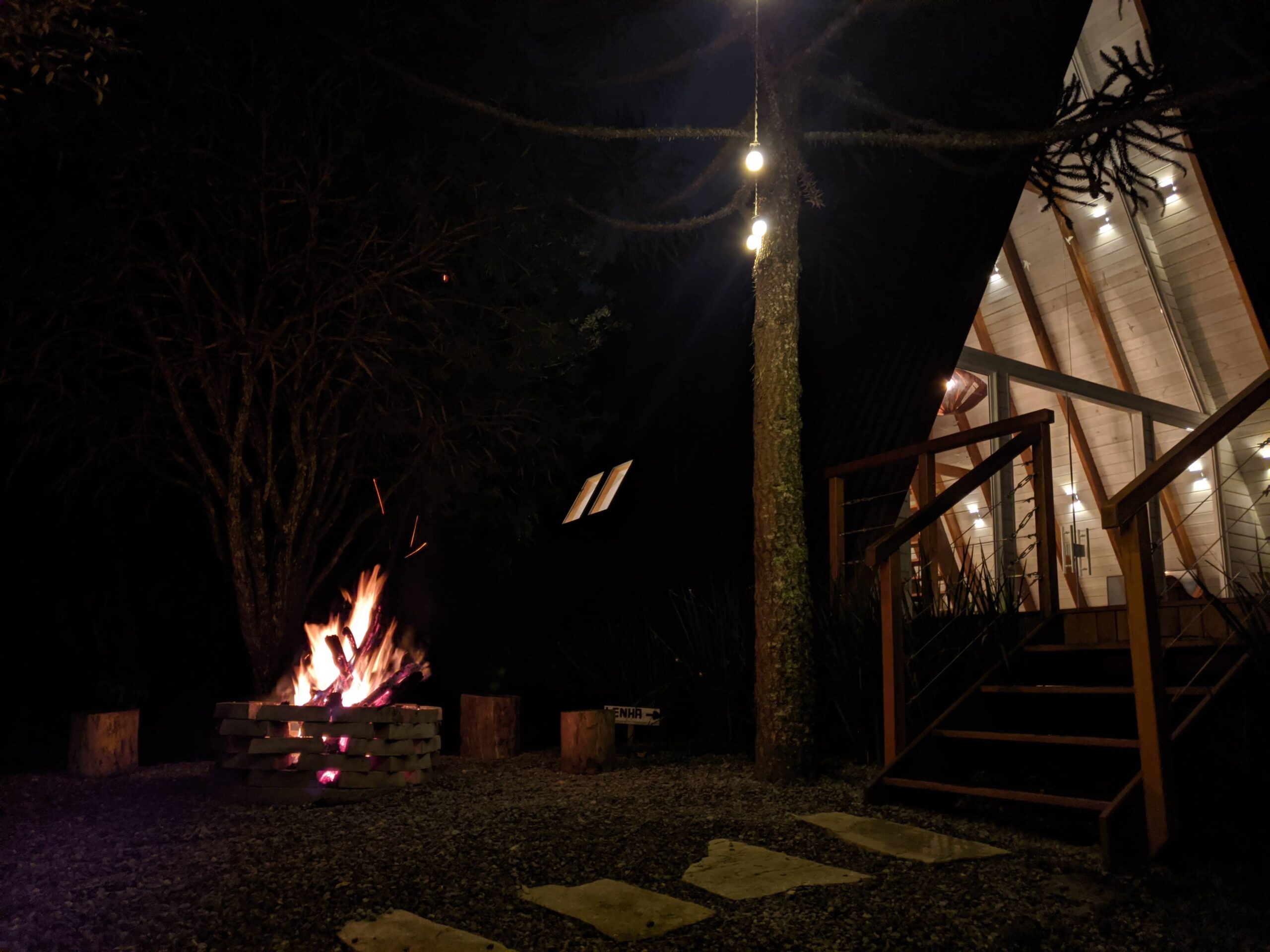 Vista noturna do exterior de uma cabana perto de Curitiba com teto em forma de A, com luzes penduradas em uma corda que se estendem até a vegetação escura. Uma fogueira acesa fornece uma fonte de luz e calor na área ao ar livre, com bancos de tronco de árvores ao redor para sentar.