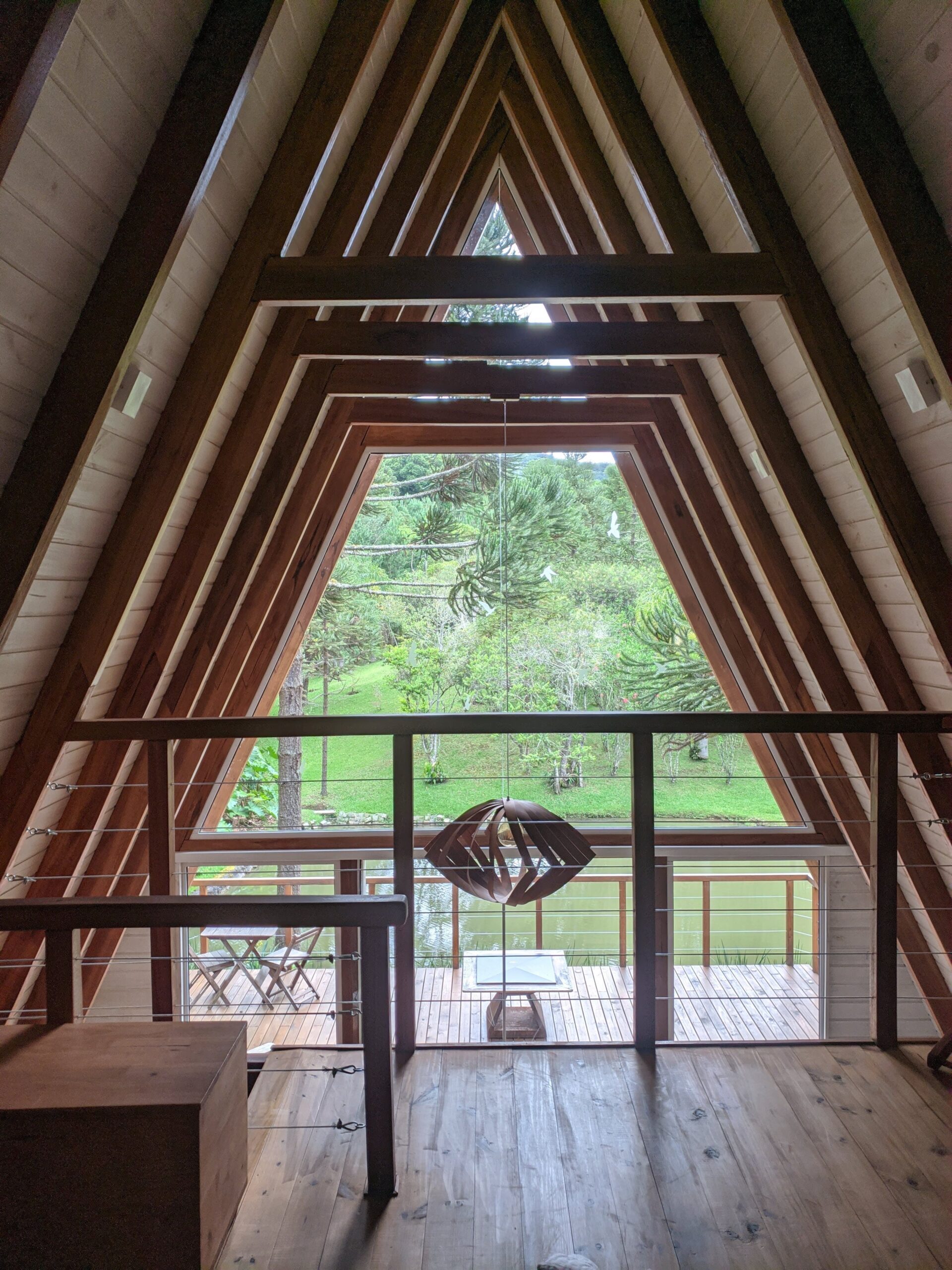 Visão interna da cabana perto de Curitiba, olhando para fora através de uma grande janela triangular que oferece uma vista ampla da natureza ao redor. O design interior em madeira cria um ambiente rústico e acolhedor.