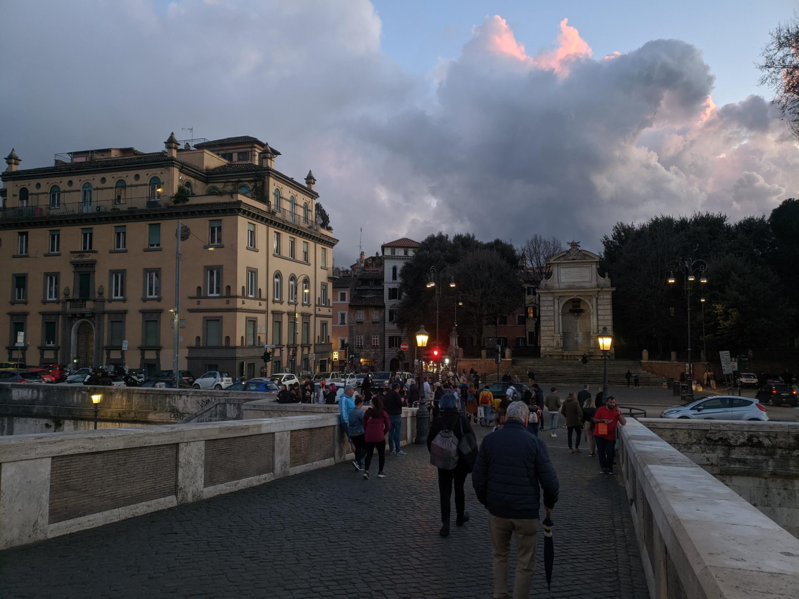 Vista do entardecer ao cruzar a Ponte Sisto em Roma, com pedestres caminhando, o céu dramático ao fundo com nuvens iluminadas pelo sol poente, e a arquitetura histórica emoldurando a cena