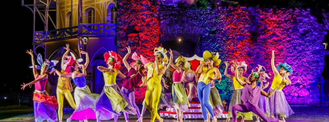 Dançarinos coloridos apresentam uma performance vibrante e energética em frente a uma estrutura iluminada, parte das celebrações de Natal em Curitiba, vestindo fantasias festivas que acrescentam um toque mágico à noite.
