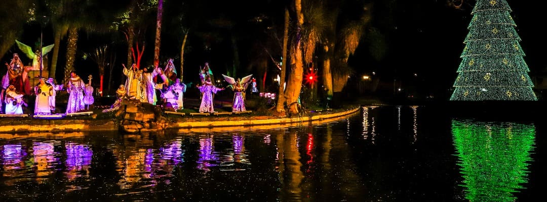 Uma cena encantadora mostra atores em trajes natalinos performáticos ao longo da margem de um lago, com uma grande árvore de Natal iluminada ao fundo, refletindo sua luz sobre as águas tranquilas, como parte dos eventos de Natal em Curitiba.
