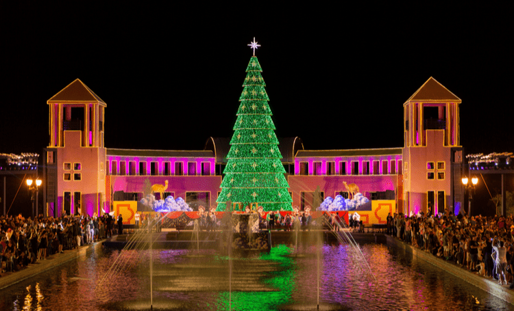 Uma imponente árvore de Natal iluminada se ergue no centro do Parque Tánguá, com a água refletindo suas luzes, enquanto os espectadores observam a decoração festiva ao redor do parque, destacando a arquitetura simétrica dos edifícios ao fundo.