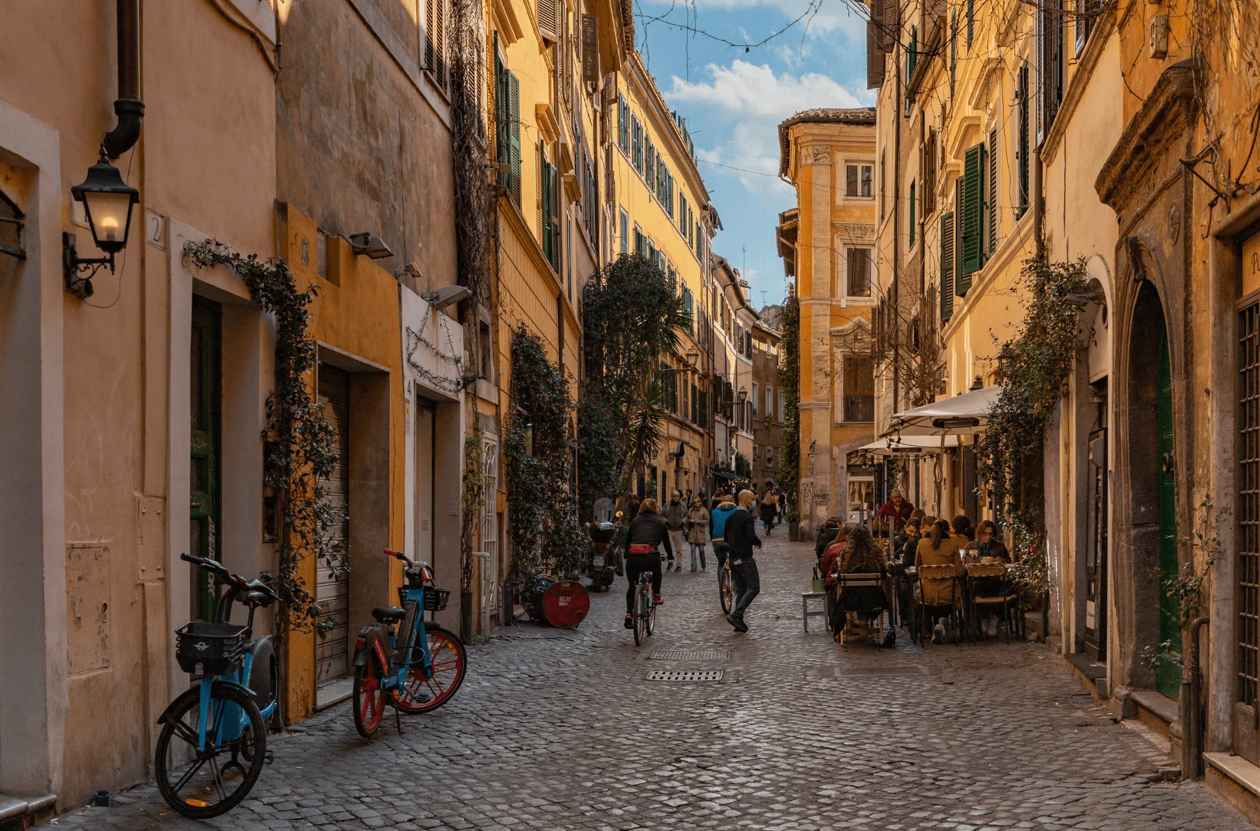 Rua vibrante e pitoresca de Trastevere, com bicicletas coloridas estacionadas, pessoas caminhando e se reunindo em um café ao ar livre, sob a luz dourada do sol que filtra pelas folhagens e edifícios históricos