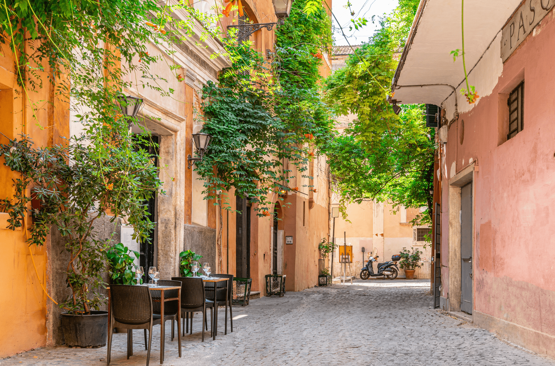 Uma tranquila rua de paralelepípedos em Trastevere, Roma, com mesas de um café ao ar livre dispostas ao lado de plantas exuberantes e fachadas de edifícios em tons quentes de amarelo e terracota