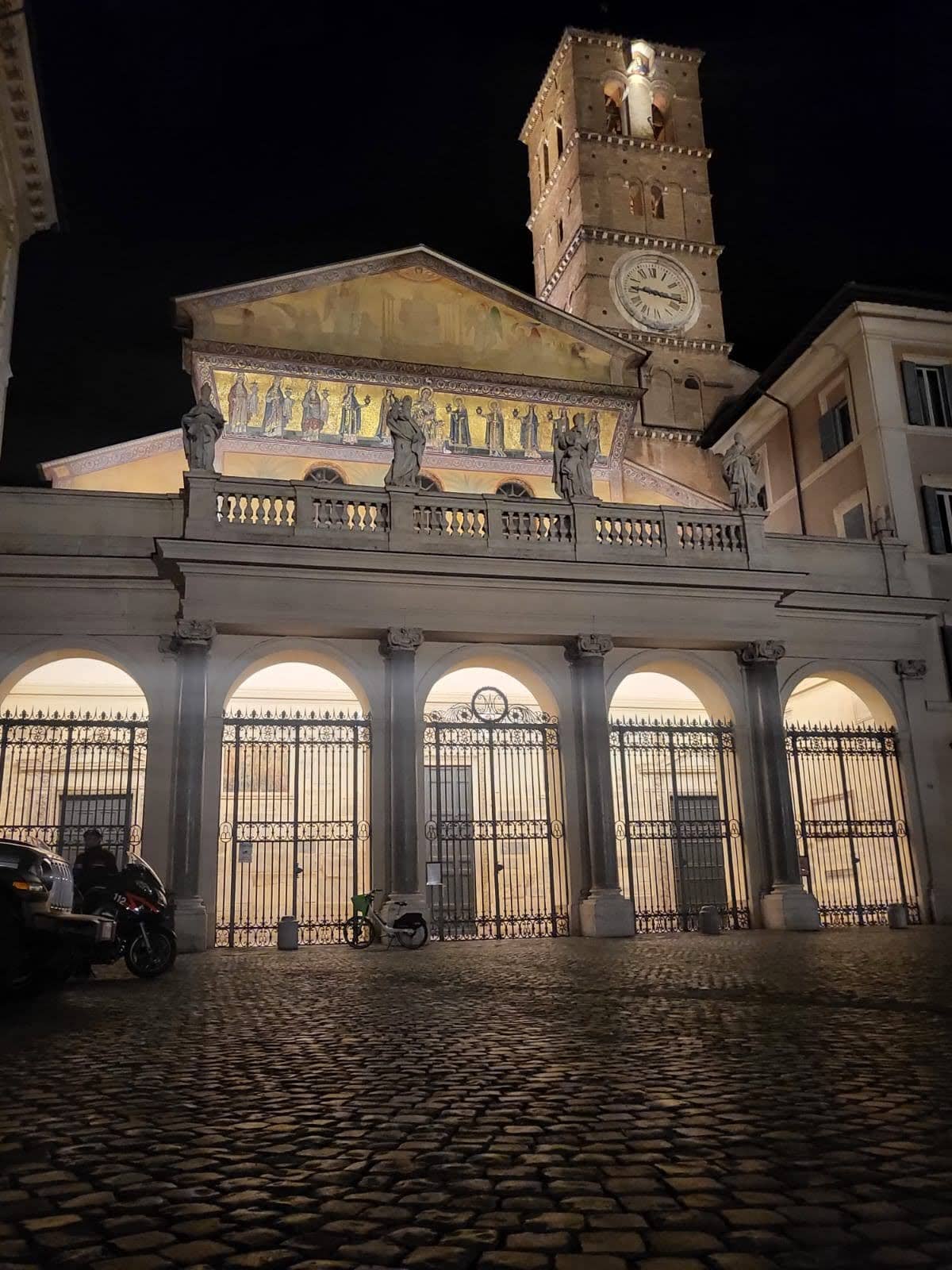 Vista noturna da fachada iluminada e da torre do sino da Basílica de Santa Maria em Trastevere, Roma, com estátuas clássicas em primeiro plano, destacando os mosaicos dourados intrincados e a arquitetura histórica da igreja.