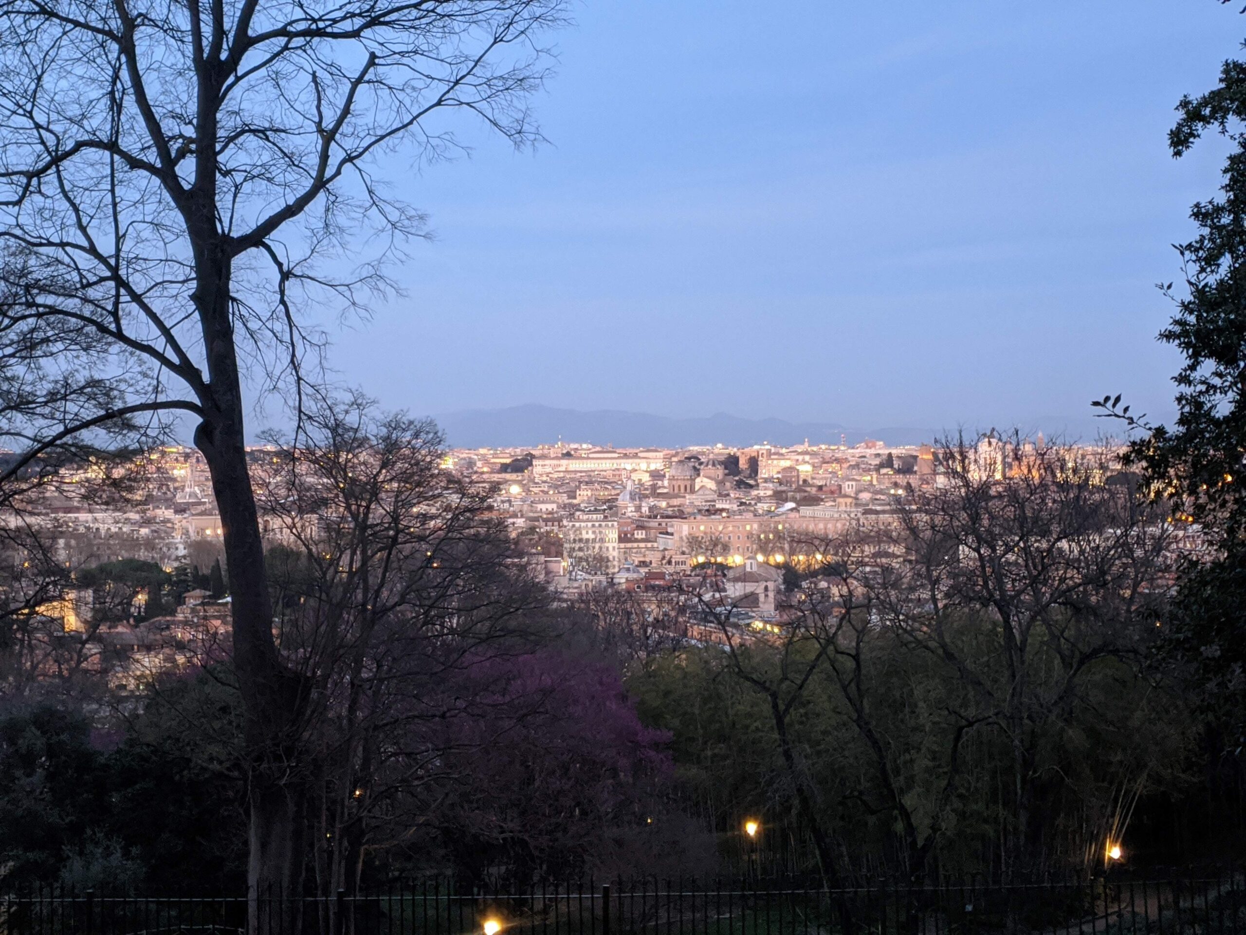 Vista panorâmica do crepúsculo sobre Roma, vista de um parque elevado com árvores desfolhadas em primeiro plano e luzes da cidade começando a brilhar sob um céu azul degradê
