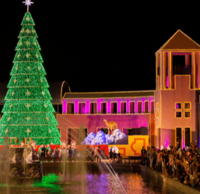 Uma imponente árvore de Natal iluminada se ergue no centro do Parque Tánguá, com a água refletindo suas luzes, enquanto os espectadores observam a decoração festiva ao redor do parque, destacando a arquitetura simétrica dos edifícios ao fundo.