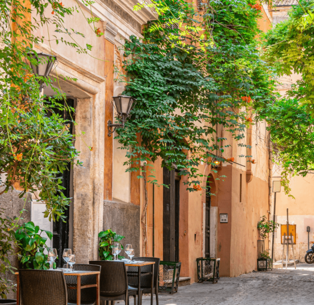 Uma tranquila rua de paralelepípedos em Trastevere, Roma, com mesas de um café ao ar livre dispostas ao lado de plantas exuberantes e fachadas de edifícios em tons quentes de amarelo e terracota